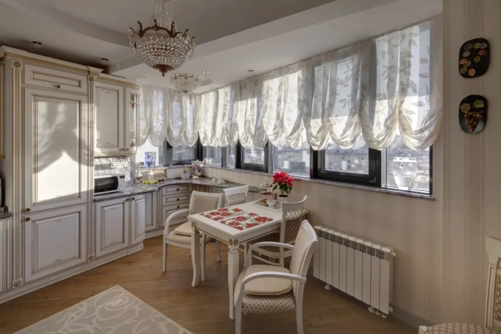 Cum Să Alegi Perdelele Potrivite pentru Fiecare Cameră: Ghidul pentru Living, Dormitor și Bucătărie Bucătărie clasică luminoasă, amenajată cu mobilă albă elegantă, candelabre cu cristale și perdele franțuzești diafane la ferestre.