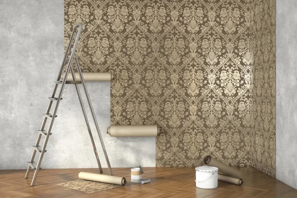 Montaj Tapet Perete: Ghidul Complet de la A la Z pentru un Finisaj Perfect Perete în curs de decorare cu tapet elegant, model damasc auriu pe fundal maro, alături de scăriță metalică, role de tapet, găleată și pensulă pentru montaj.