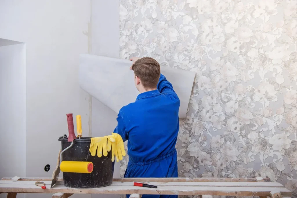 Montaj Tapet Perete: Ghidul Complet de la A la Z pentru un Finisaj Perfect Un muncitor montează tapet decorativ, având la îndemână găleată cu adeziv, mistrie, cutter, rolă de aplicare și mănuși de protecție.